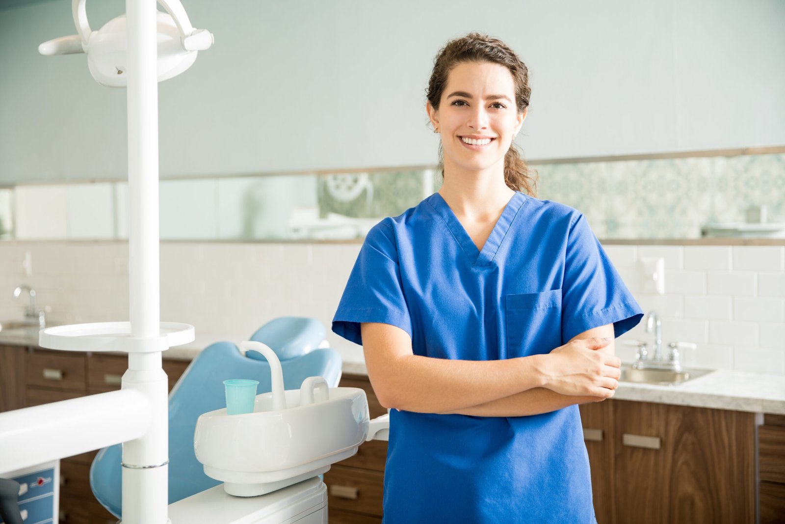 Portrait of smiling female dentist in scrubs standing with arms crossed at clinic