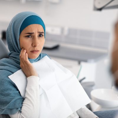 Teeth Problems. Muslim Female Patient In Dentist Chair Showing Aching Tooth To Doctor, Upset Islamic Woman In Hijab Touching Sore Cheek During Treatment In Modern Stomatologic Clinic, Closeup